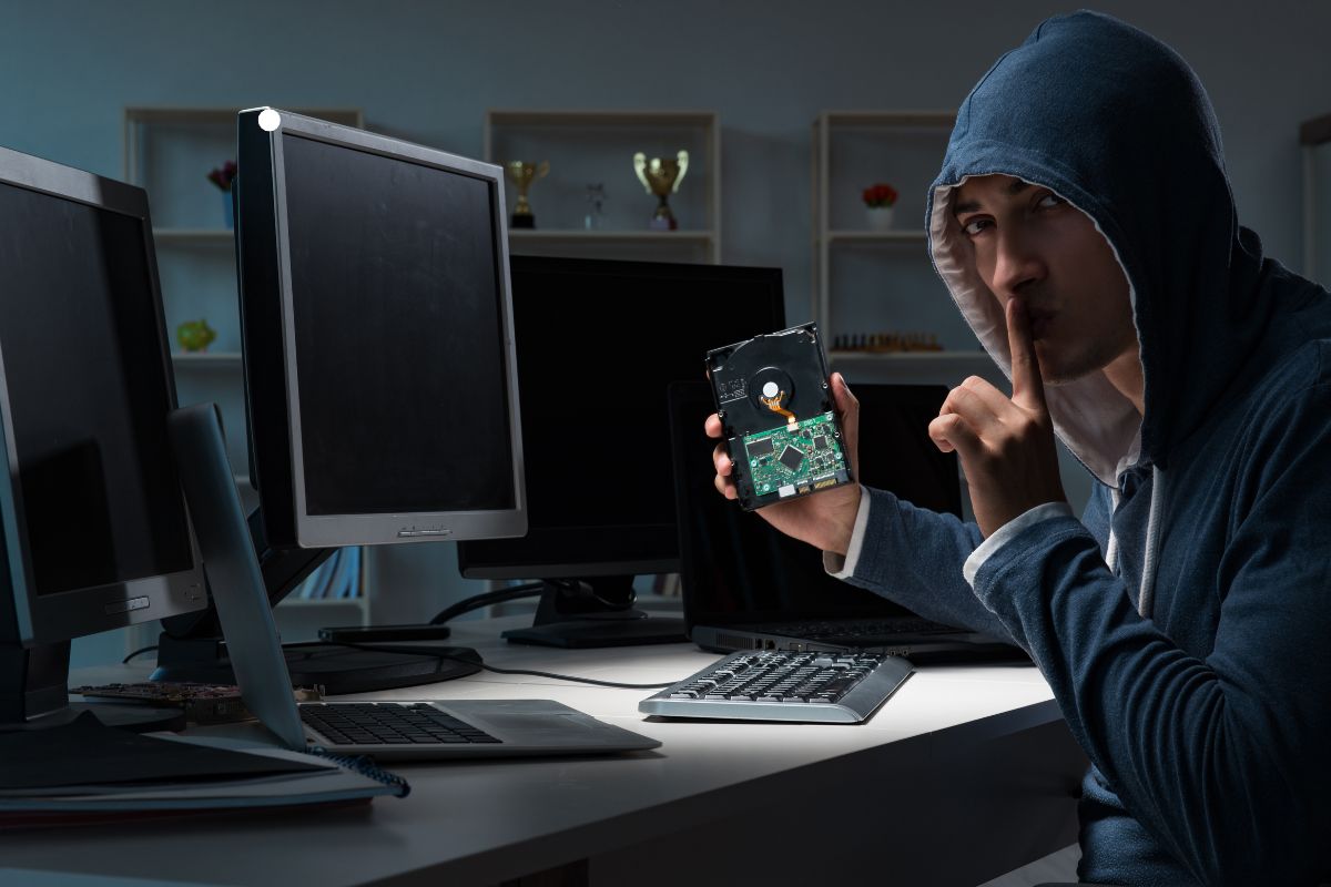 A hooded figure silently holds a disassembled hard drive, surrounded by multiple dark computer monitors on a desk.