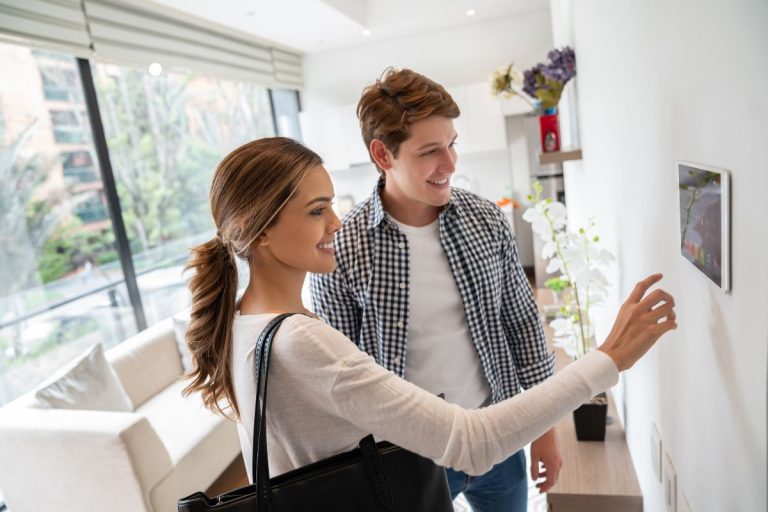 A woman in a white top operates a wall-mounted tablet as a man in a checked shirt observes, in a bright, modern living space.