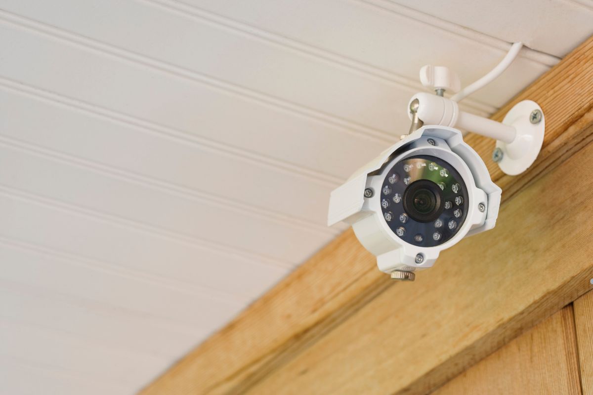 A white security camera mounted on a wooden wall, with a white ceiling and a focus on its lens and LED indicators.