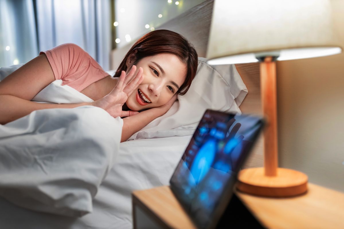 A person lies in bed with a laptop on the nightstand, surrounded by soft lighting and cozy blankets.
